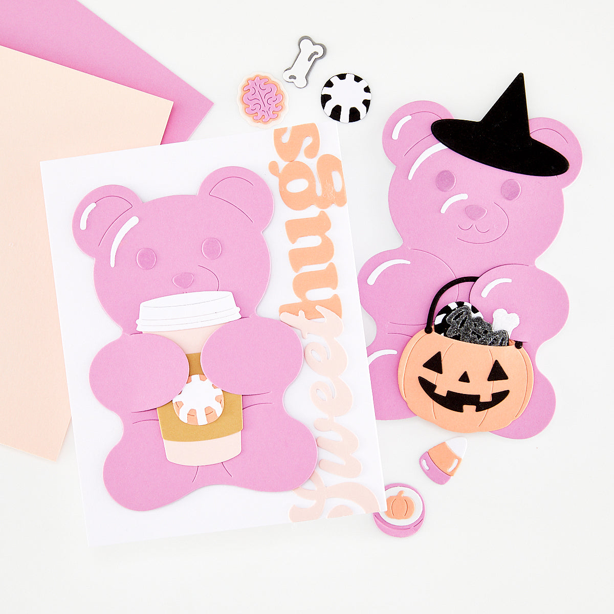 A styled flat-lay of pink bear-shaped craft cutouts and handmade card on a white background, including a bear holding a coffee cup, a bear wearing a witch hat, a jack-o'-lantern bucket, small decorative pieces, and a vertical 'Sweethugs' word.
