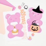 A styled flat-lay of pink bear-shaped craft cutouts and handmade card on a white background, including a bear holding a coffee cup, a bear wearing a witch hat, a jack-o'-lantern bucket, small decorative pieces, and a vertical 'Sweethugs' word.