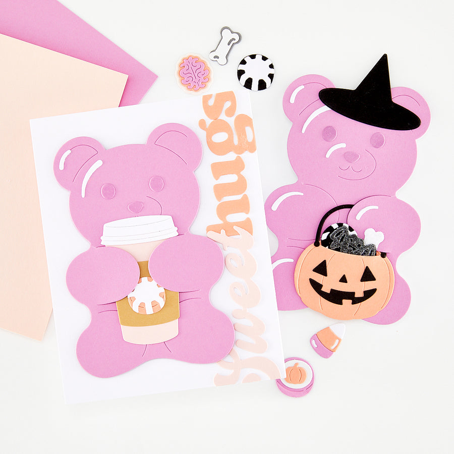 A styled flat-lay of pink bear-shaped craft cutouts and handmade card on a white background, including a bear holding a coffee cup, a bear wearing a witch hat, a jack-o'-lantern bucket, small decorative pieces, and a vertical 'Sweethugs' word.