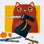 A handmade autumn-themed greeting card of a brown cat holding an Amazon-branded card, styled with fall decorations including pumpkins, leaves, and text stickers on a white background.