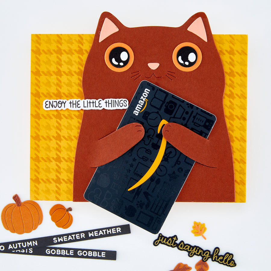 A handmade autumn-themed greeting card of a brown cat holding an Amazon-branded card, styled with fall decorations including pumpkins, leaves, and text stickers on a white background.