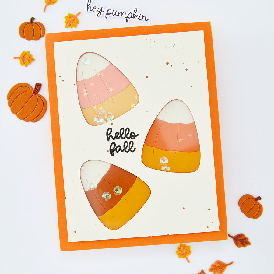 A handmade autumn greeting card with an orange border and three candy corn cutouts showing pastel and warm orange layers. The card says 'hello fall' in black script on a cream background, styled with small pumpkins and leaves on a white surface.