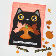 A handmade autumn-themed greeting card featuring a black cat holding a pumpkin with layered fall leaves, displayed on a white surface with decorative pumpkins and leaves around it.