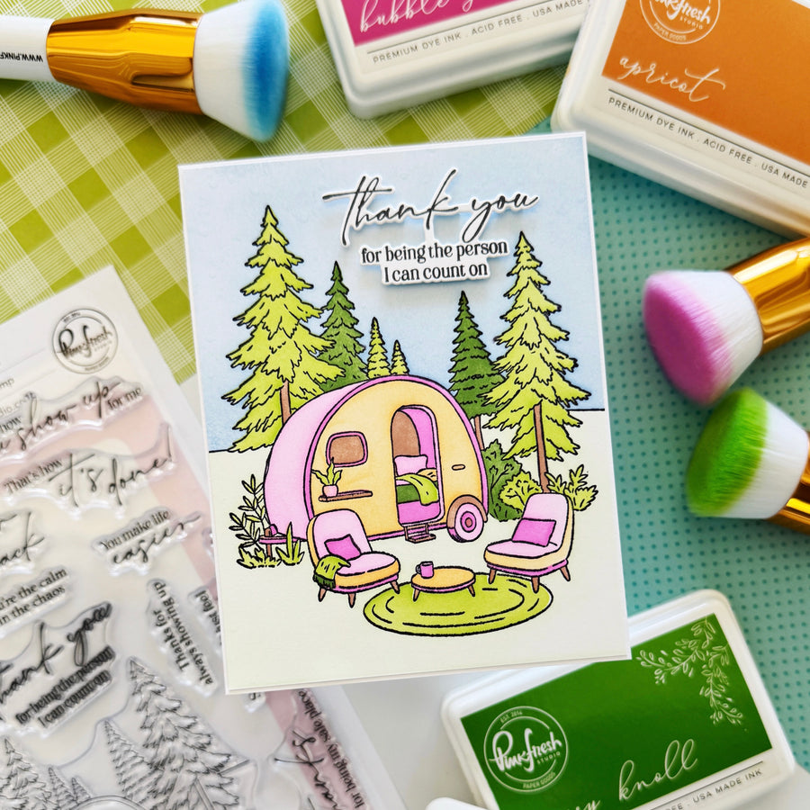 A handmade greeting card featuring a retro pink and yellow camper trailer with camping chairs and trees, surrounded by craft supplies including stamp sheets, ink pads, and makeup brushes on a green plaid and teal background.