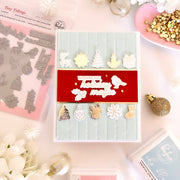 A flat-lay of a handmade holiday card with layered white and gold die-cut embellishments, shown alongside stamping tools and craft supplies on a light pink surface.