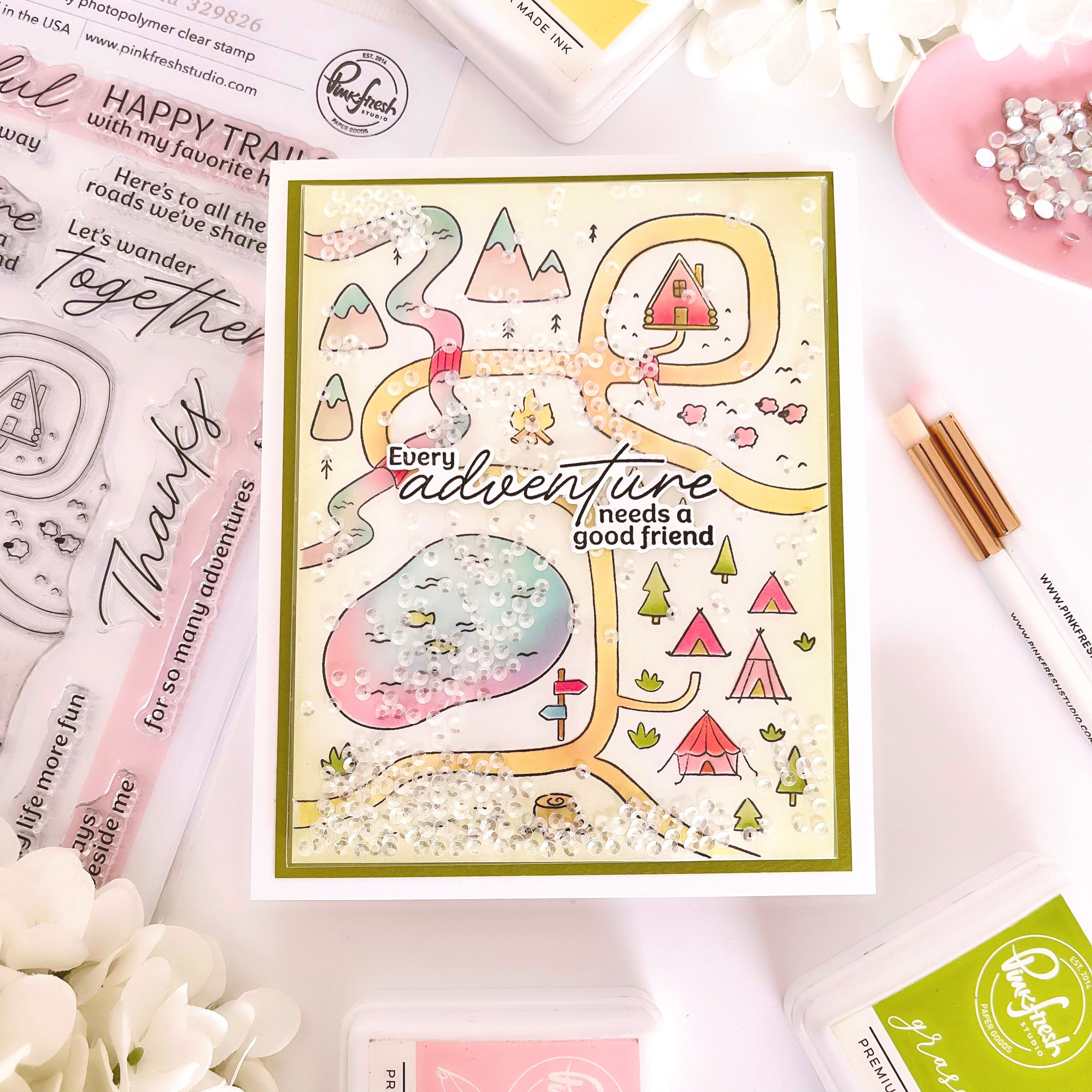 A handmade glittery camping map card with a green border, surrounded by crafting supplies including Ranger brand stamps, an ink pad, a white pen, and a pink dish with rhinestones on a white background.