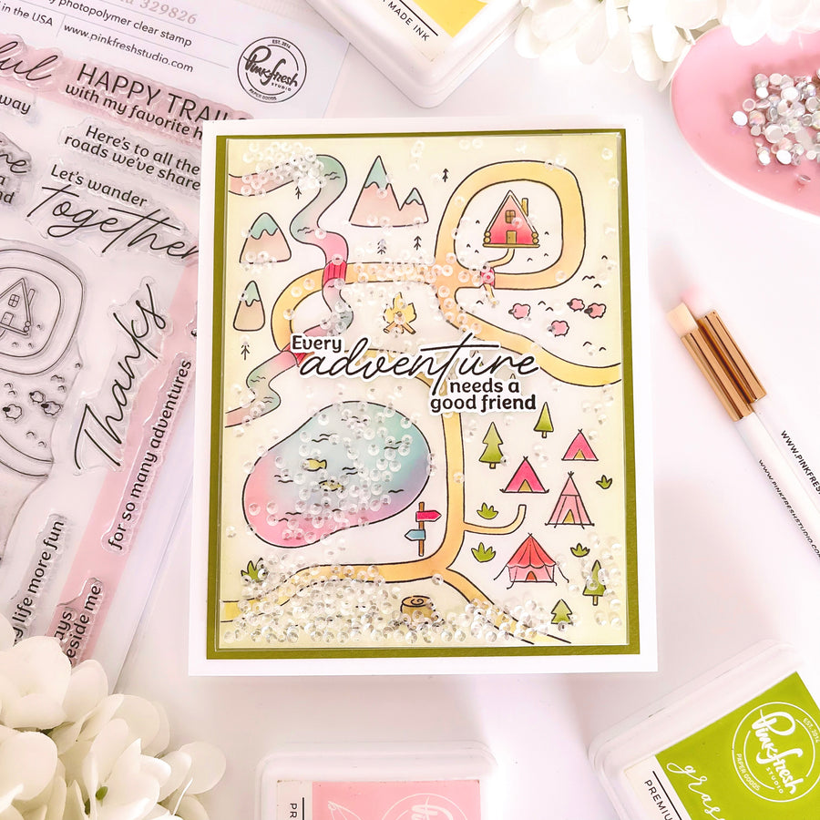 A handmade glittery camping map card with a green border, surrounded by crafting supplies including Ranger brand stamps, an ink pad, a white pen, and a pink dish with rhinestones on a white background.