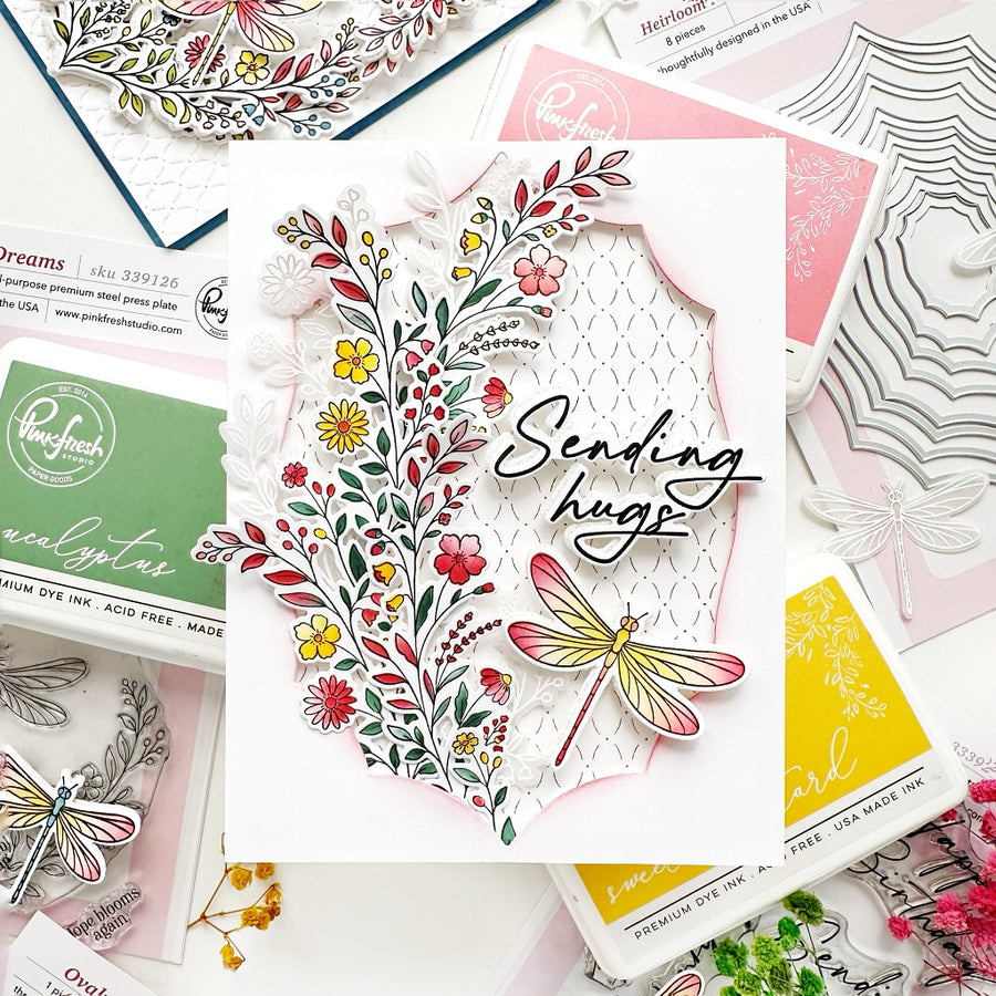 A handmade layered card with a floral cutout design and a dragonfly, surrounded by craft stamp and die packaging on a white background.