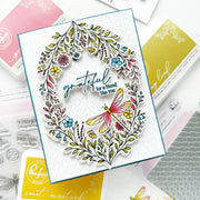 A handmade greeting card featuring a colorful floral wreath and a dragonfly, with the sentiment 'grateful for a friend like you' in blue script. The card is displayed on a flat-lay arrangement of crafting supplies, including RangerFleet stamps and ink pads.