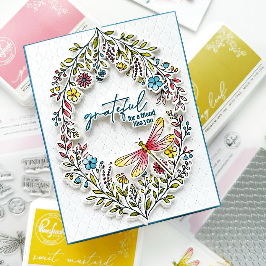 A handmade greeting card featuring a colorful floral wreath and a dragonfly, with the sentiment 'grateful for a friend like you' in blue script. The card is displayed on a flat-lay arrangement of crafting supplies, including RangerFleet stamps and ink pads.