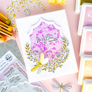 A handmade layered card featuring a central shield-shaped design with a purple glittery pink background, the words 'brighter days away,' and a yellow dragonfly surrounded by delicate floral illustrations. The card is styled on a pink background with craft supplies, including ink pads, stamps, and paintbrushes.