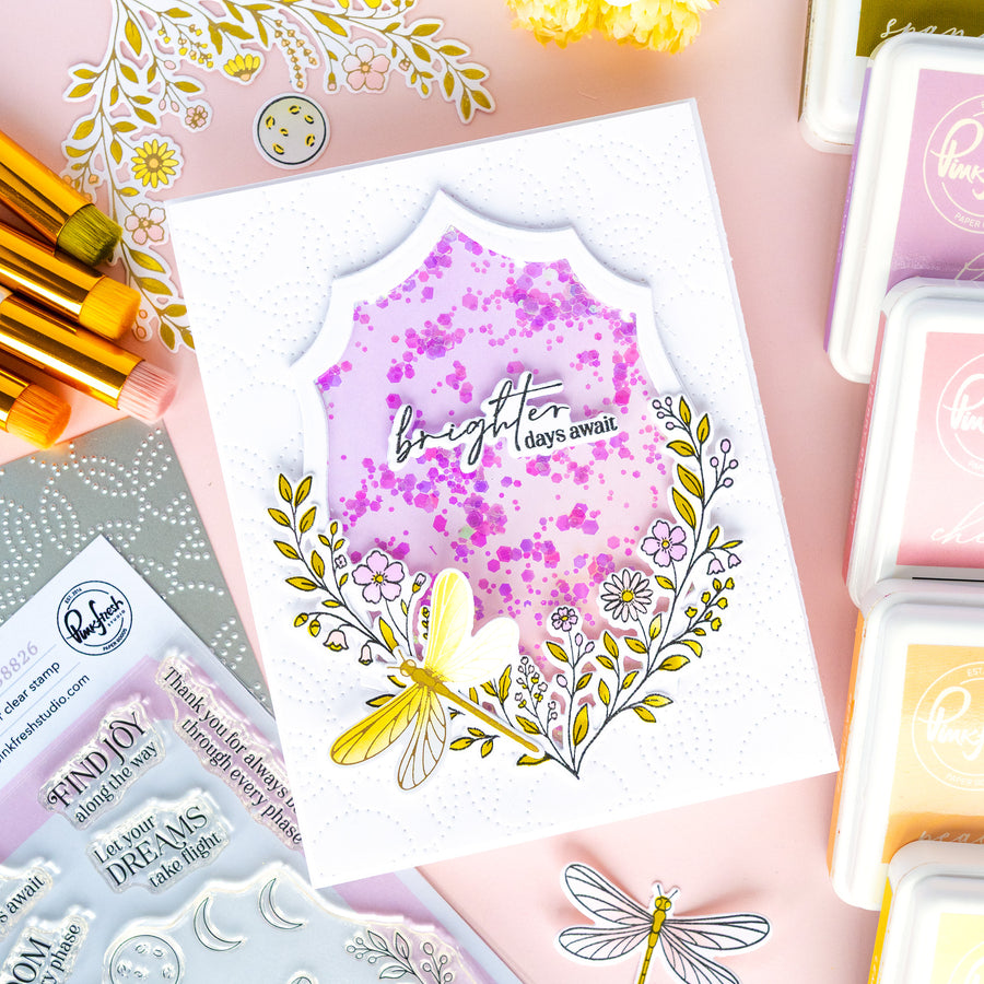 A handmade layered card featuring a central shield-shaped design with a purple glittery pink background, the words 'brighter days away,' and a yellow dragonfly surrounded by delicate floral illustrations. The card is styled on a pink background with craft supplies, including ink pads, stamps, and paintbrushes.