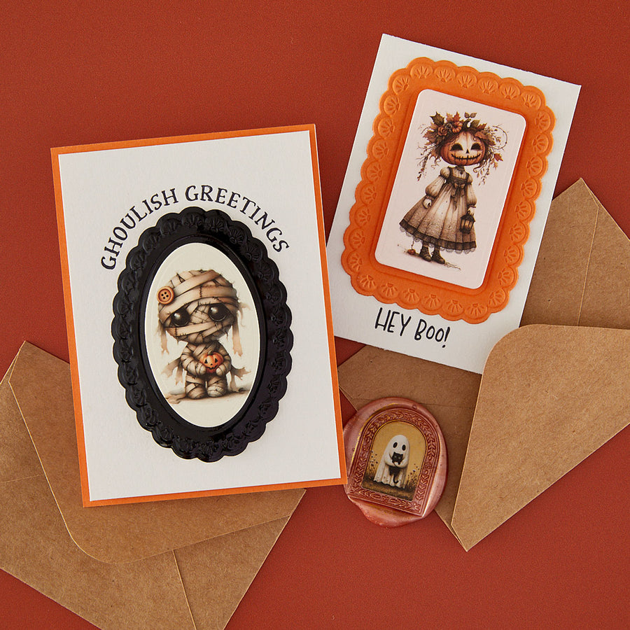 Two Halloween greeting cards with decorative frames and illustrations, shown with brown envelopes on a warm red-orange background. One card reads 'Ghoulish Greetings' with a mummy illustration, and the other reads 'HEY BOO!' with a ghostly figure. Uses Spellbinders Tricks & Treats Rub-On Sheets
