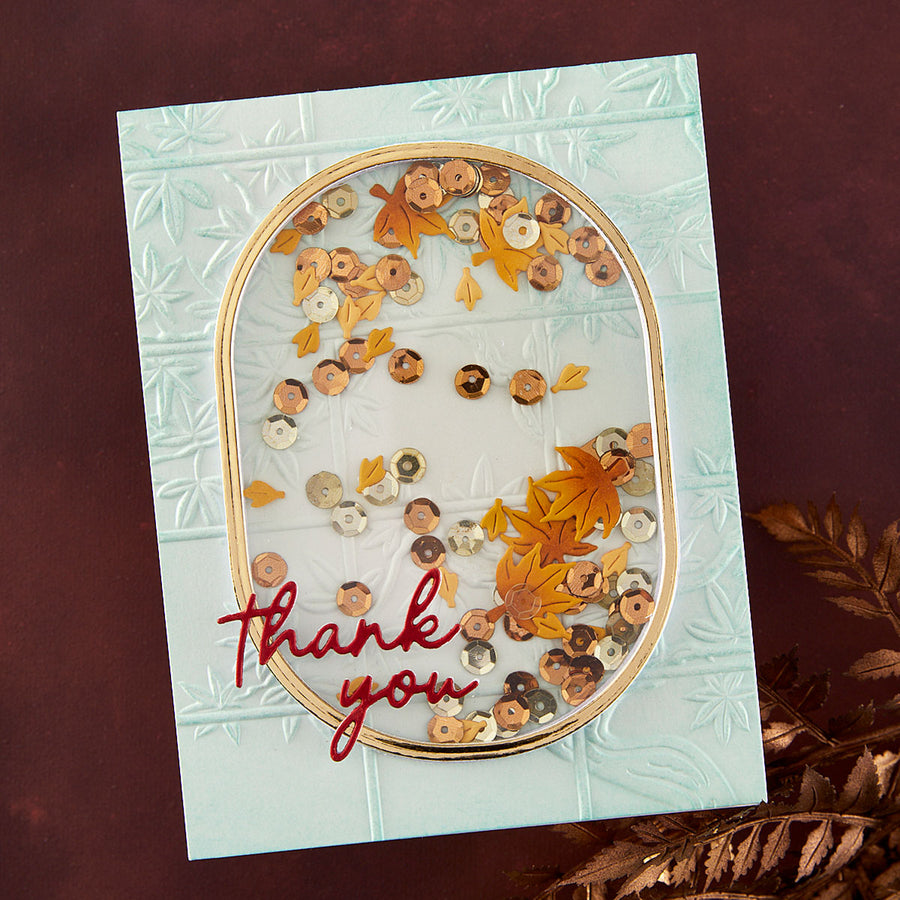 A handmade square greeting card with a light aqua embossed background, featuring a central oval gold frame filled with autumn sequins and dried orange and brown leaves. The card has the words 'thank you' in red script at the bottom left and is photographed on a dark reddish-brown surface with dried foliage in the corner.