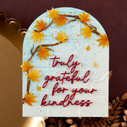 A handmade autumn-themed card featuring an arched white background with embossed floral details, decorated with 3D yellow maple leaves on brown branches and the phrase 'truly grateful for your kindness' in red script.