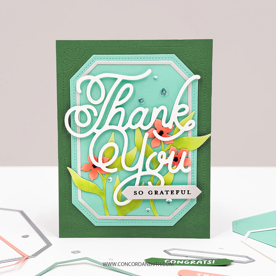 A handmade layered greeting card with a dark green base, a mint green inner panel, white script reading 'Thank You', and floral accents in coral and green, displayed on a white surface with crafting tools nearby.