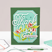 A handmade layered greeting card with a dark green base, a mint green inner panel, white script reading 'Thank You', and floral accents in coral and green, displayed on a white surface with crafting tools nearby.