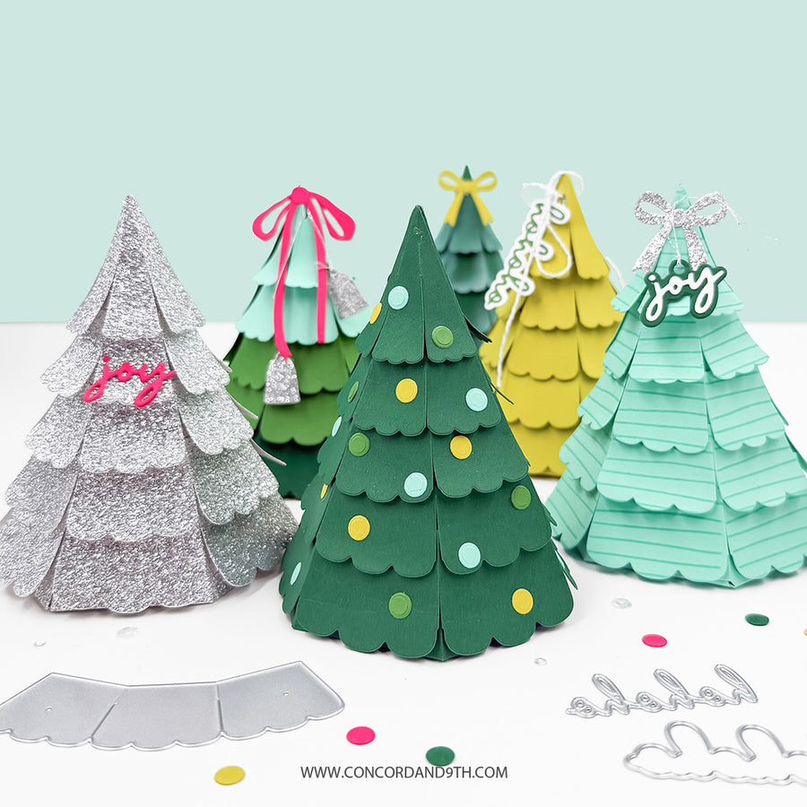 A set of Christmas tree-shaped paper craft ornaments in silver, dark green, yellow, and mint green, displayed with matching silver paper dies on a white surface against a light mint green background. One ornament has a pink bow and the mint green one has a silver 'Joy' script topper.