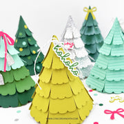 A set of colorful, tiered paper Christmas tree ornaments displayed on a white surface with festive confetti. The central ornament is bright yellow with a white hanging string and a green 'Merry Christmas' banner.