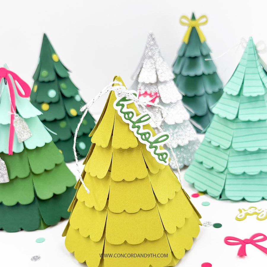 A set of colorful, tiered paper Christmas tree ornaments displayed on a white surface with festive confetti. The central ornament is bright yellow with a white hanging string and a green 'Merry Christmas' banner.