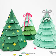 Three handmade layered paper Christmas tree ornaments in different green shades, decorated with colorful circular accents, a pink ribbon, and a silver bow with a 'joy' hanging ornament, displayed on a white surface.