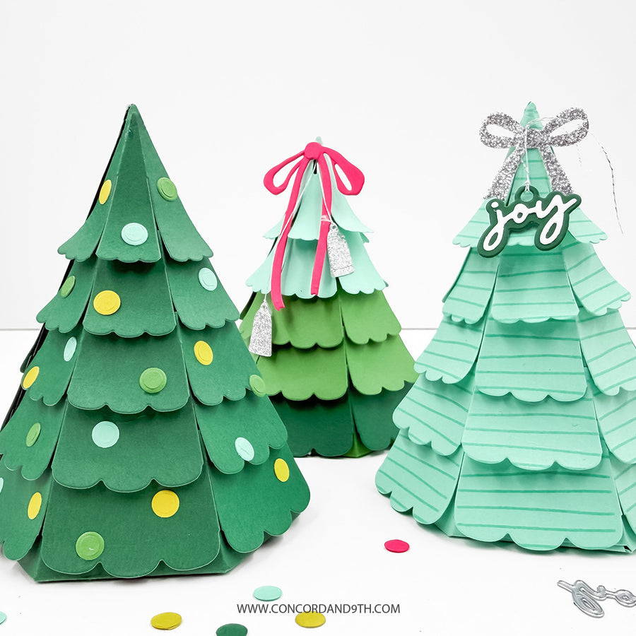 Three handmade layered paper Christmas tree ornaments in different green shades, decorated with colorful circular accents, a pink ribbon, and a silver bow with a 'joy' hanging ornament, displayed on a white surface.