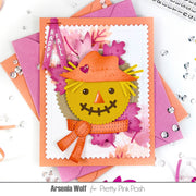 A handmade autumn-themed greeting card featuring a smiling scarecrow face with a yellow head, orange hat, and orange scarf, layered with pink and orange leaves. The card is styled on top of pink and orange envelopes with scattered rhinestones on a white background.