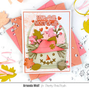 A handmade autumn-themed greeting card featuring a smiling scarecrow with a pink hat and layered fall leaves, surrounded by pink and peach envelopes, craft embellishments, and decorative leaves on a white background. Text at the bottom reads 'Arsenia Wolf for Pretty Pink Posh'.