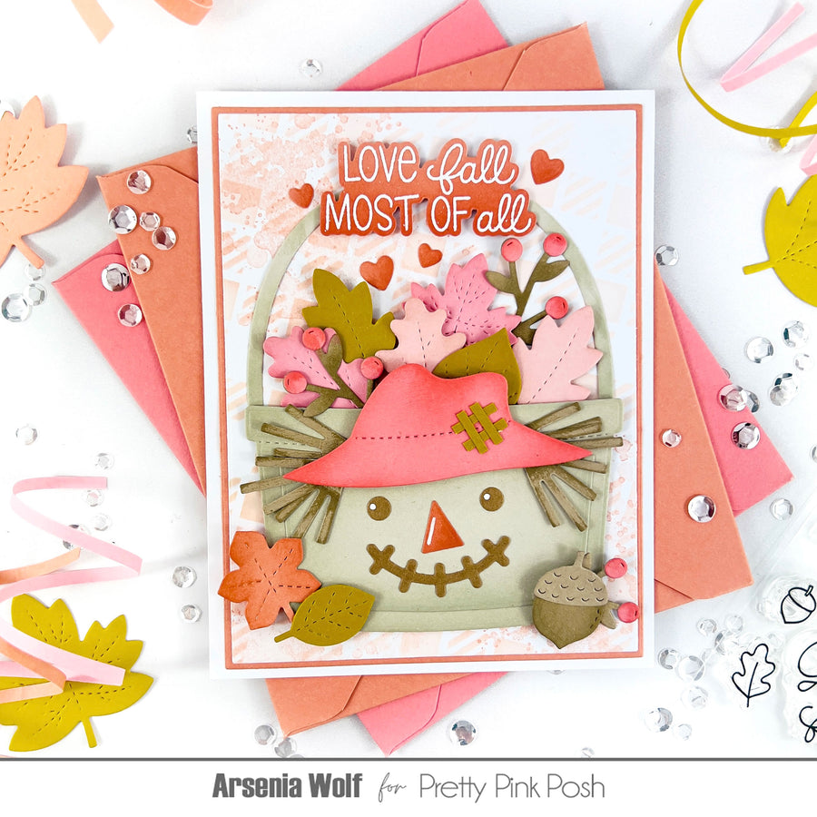 A handmade autumn-themed greeting card featuring a smiling scarecrow with a pink hat and layered fall leaves, surrounded by pink and peach envelopes, craft embellishments, and decorative leaves on a white background. Text at the bottom reads 'Arsenia Wolf for Pretty Pink Posh'.