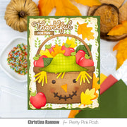A handmade autumn-themed greeting card featuring a smiling scarecrow basket with red apples and fall leaves, styled with pumpkins, autumn leaves, and craft supplies on a white surface. Text at the bottom reads 'Christina Rannow for Pretty Pink Posh'.