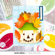 A handmade autumn scarecrow card featuring a cream-colored scarecrow face with a green hat and orange, yellow, and brown paper leaves as hair, set on a light blue patterned background. The card is displayed in a flat-lay arrangement with Stampin' Up! ink pads and other crafting supplies blurred in the background.