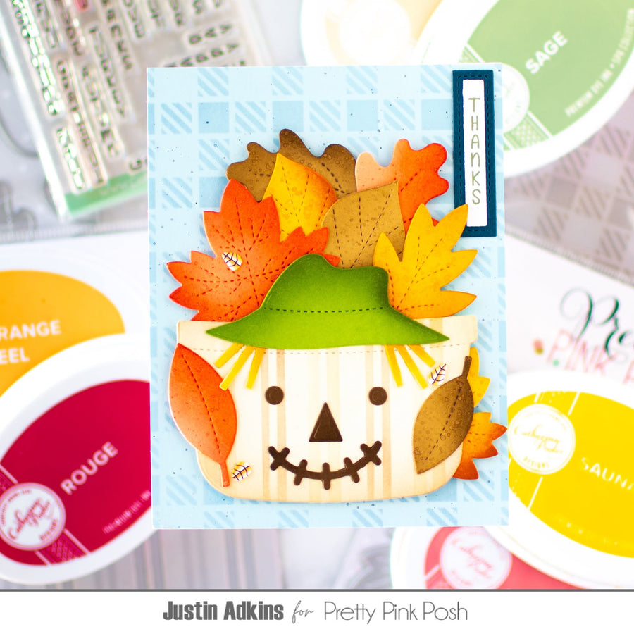 A handmade autumn scarecrow card featuring a cream-colored scarecrow face with a green hat and orange, yellow, and brown paper leaves as hair, set on a light blue patterned background. The card is displayed in a flat-lay arrangement with Stampin' Up! ink pads and other crafting supplies blurred in the background.
