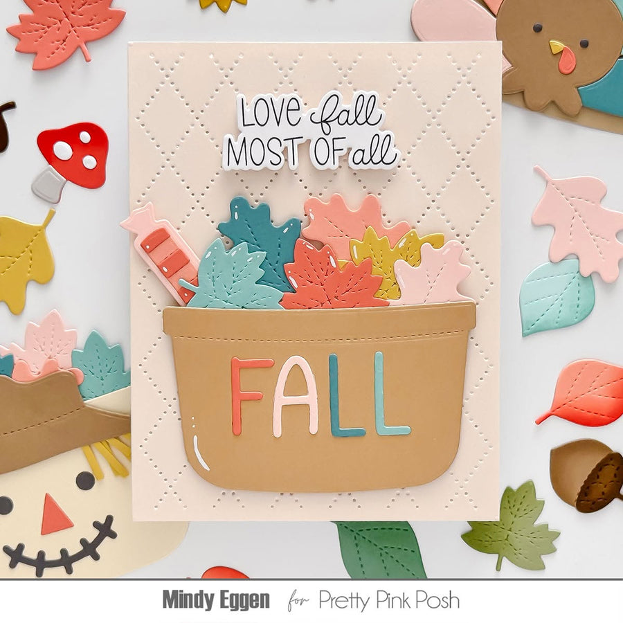 A handmade autumn-themed card featuring a tan basket filled with colorful die-cut fall leaves and a small red-orange pumpkin, with the words 'LOVE fall MOST of all' above and 'FALL' spelled out in letters on the front. The card is surrounded by scattered fall-themed paper decorations, including leaves, acorns, a scarecrow, and a turkey.