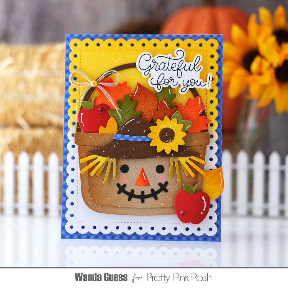 A handmade autumn-themed greeting card featuring a smiling scarecrow face inside a basket filled with colorful fall leaves, apples, and a sunflower. The card has a scalloped blue border and the phrase 'Grateful for you!' in white script.