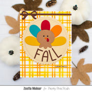 A handmade Thanksgiving greeting card featuring a turkey made from layered paper feathers in red, teal, orange, and yellow, with the word 'FALL' and 'GREETINGS' printed on it. The card has a yellow and white plaid border and a brown ribbon accent at the top.