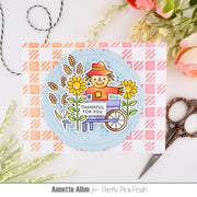 A handmade greeting card featuring a smiling scarecrow in a wheelbarrow surrounded by sunflowers and wheat, with the text 'THANKFUL FOR YOU'. The card is layered over a pink and white gingham patterned sheet.