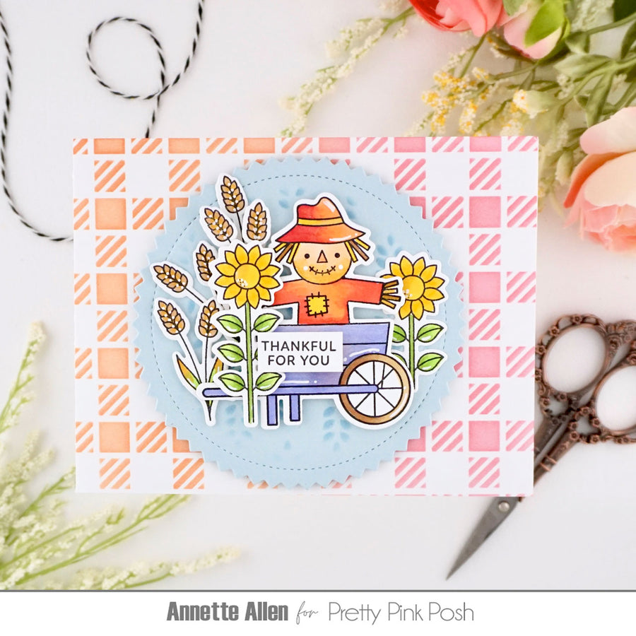 A handmade greeting card featuring a smiling scarecrow in a wheelbarrow surrounded by sunflowers and wheat, with the text 'THANKFUL FOR YOU'. The card is layered over a pink and white gingham patterned sheet.