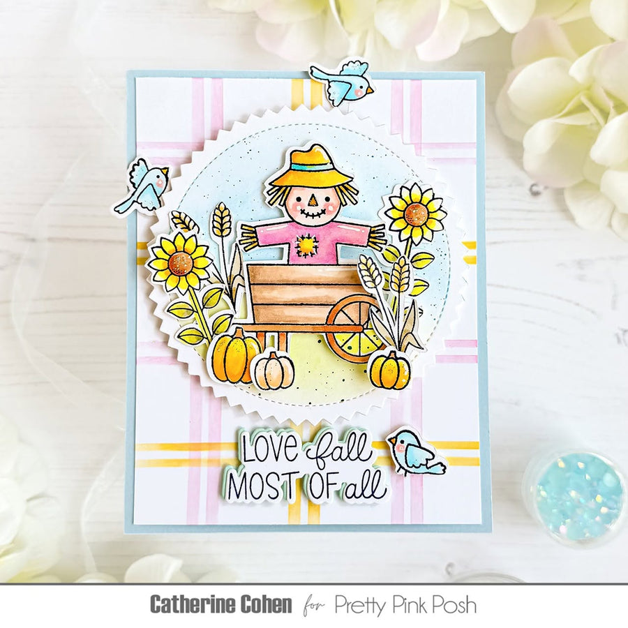 A handmade autumn-themed greeting card featuring a cartoon scarecrow in a wagon with pumpkins, sunflowers, birds, and the text 'LOVE fall MOST OF all'. The card is displayed on a white wooden surface with soft white flowers in the background.