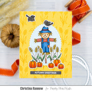 A handmade autumn-themed greeting card featuring a scarecrow, pumpkins, wheat, and birds on a yellow patterned background, styled with orange ribbons and dried grass in a flat-lay arrangement. Text at the bottom reads 'Christina Rannow for Pretty Pink Posh'.
