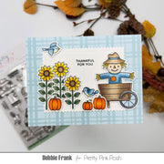 A handmade autumn-themed card featuring a scarecrow in a wagon, sunflowers, pumpkins, and blue birds on a light blue plaid background, with the text 'THANKFUL FOR YOU'. The card is displayed on a white surface with styled fall leaves and a clear stamp sheet in the background.