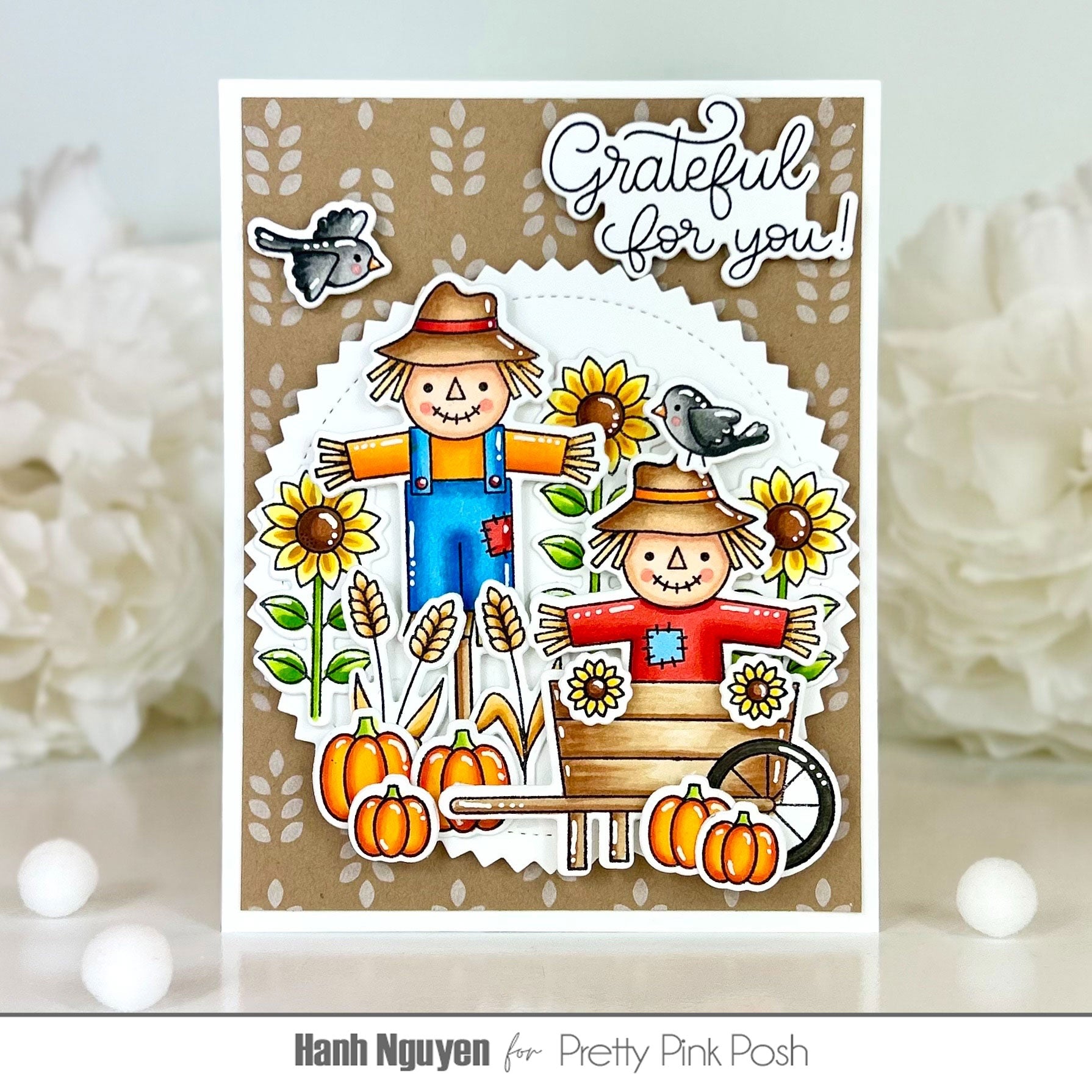 A handmade autumn-themed greeting card featuring two scarecrows, sunflowers, pumpkins, birds, and the text 'Grateful for you!' on a white and kraft paper background, displayed with blurred white flowers.