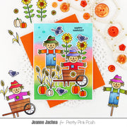 A handmade autumn-themed greeting card featuring two scarecrows, sunflowers, pumpkins, birds, and a rainbow gradient background, styled on a white surface with decorative buttons, flowers, and paper accents. The card is branded with 'Pretty Pink Posh' and designer name 'Jeanne Jachna'.