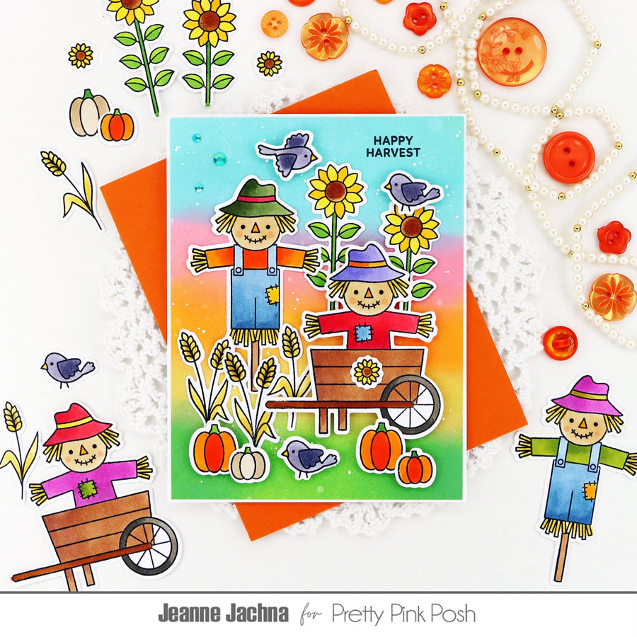 A handmade autumn-themed greeting card featuring two scarecrows, sunflowers, pumpkins, birds, and a rainbow gradient background, styled on a white surface with decorative buttons, flowers, and paper accents. The card is branded with 'Pretty Pink Posh' and designer name 'Jeanne Jachna'.