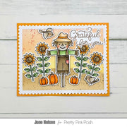 A handmade greeting card featuring a scarecrow, sunflowers, pumpkins, birds, and the text 'Grateful for you!' on a scalloped white border with an orange inner border, displayed on a light wood background.
