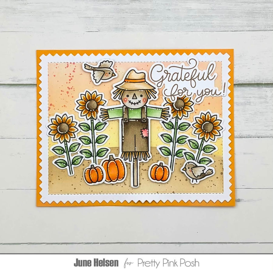 A handmade greeting card featuring a scarecrow, sunflowers, pumpkins, birds, and the text 'Grateful for you!' on a scalloped white border with an orange inner border, displayed on a light wood background.