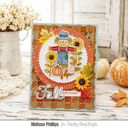 A handmade autumn-themed greeting card featuring a smiling scarecrow, sunflowers, pumpkins, and the words 'Fall Greetings' in layered scalloped paper. The card is styled on a rustic white wooden table with decorative pumpkins, a lace doily, and a white pitcher of dried flowers in the background.