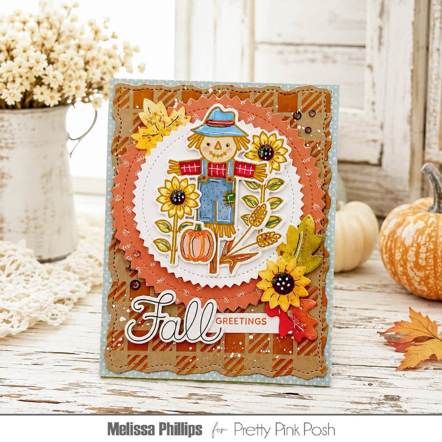 A handmade autumn-themed greeting card featuring a smiling scarecrow, sunflowers, pumpkins, and the words 'Fall Greetings' in layered scalloped paper. The card is styled on a rustic white wooden table with decorative pumpkins, a lace doily, and a white pitcher of dried flowers in the background.