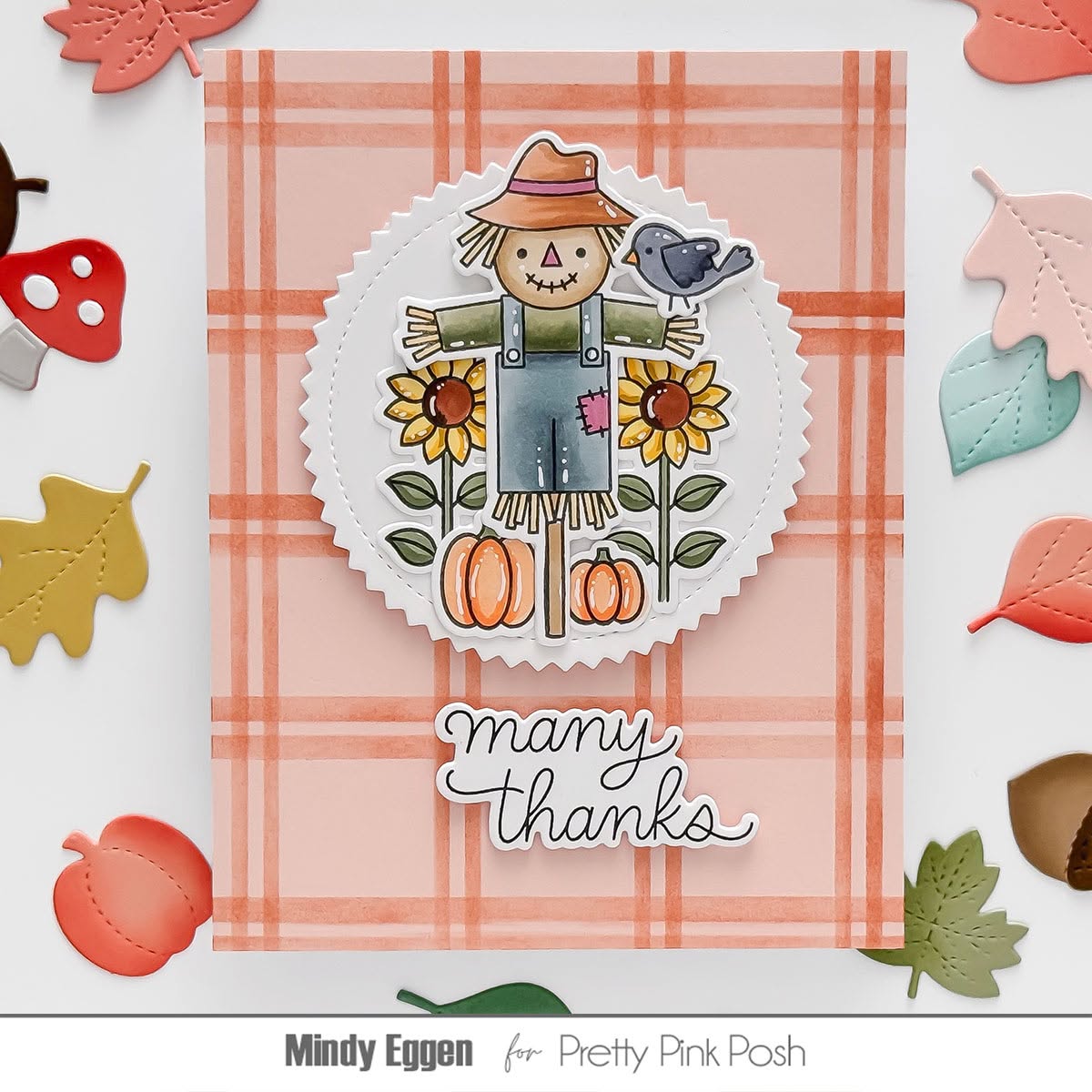 A handmade autumn-themed greeting card featuring a scalloped white circle with a die-cut scarecrow, sunflowers, pumpkins, and a bird, set on a peach plaid background. The card is layered with the phrase 'many thanks' in white script.