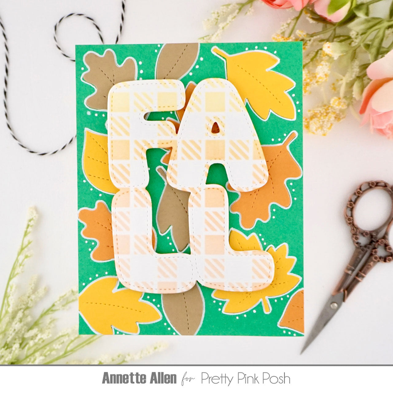 A handmade autumn-themed card featuring the word 'FALL' in layered white letters with a yellow plaid pattern, set against a green background decorated with orange, yellow, and brown leaves. The card is styled on a white surface with scissors, twine, and flowers.