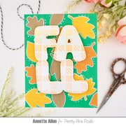 A handmade autumn-themed card featuring the word 'FALL' in layered white letters with a yellow plaid pattern, set against a green background decorated with orange, yellow, and brown leaves. The card is styled on a white surface with scissors, twine, and flowers.